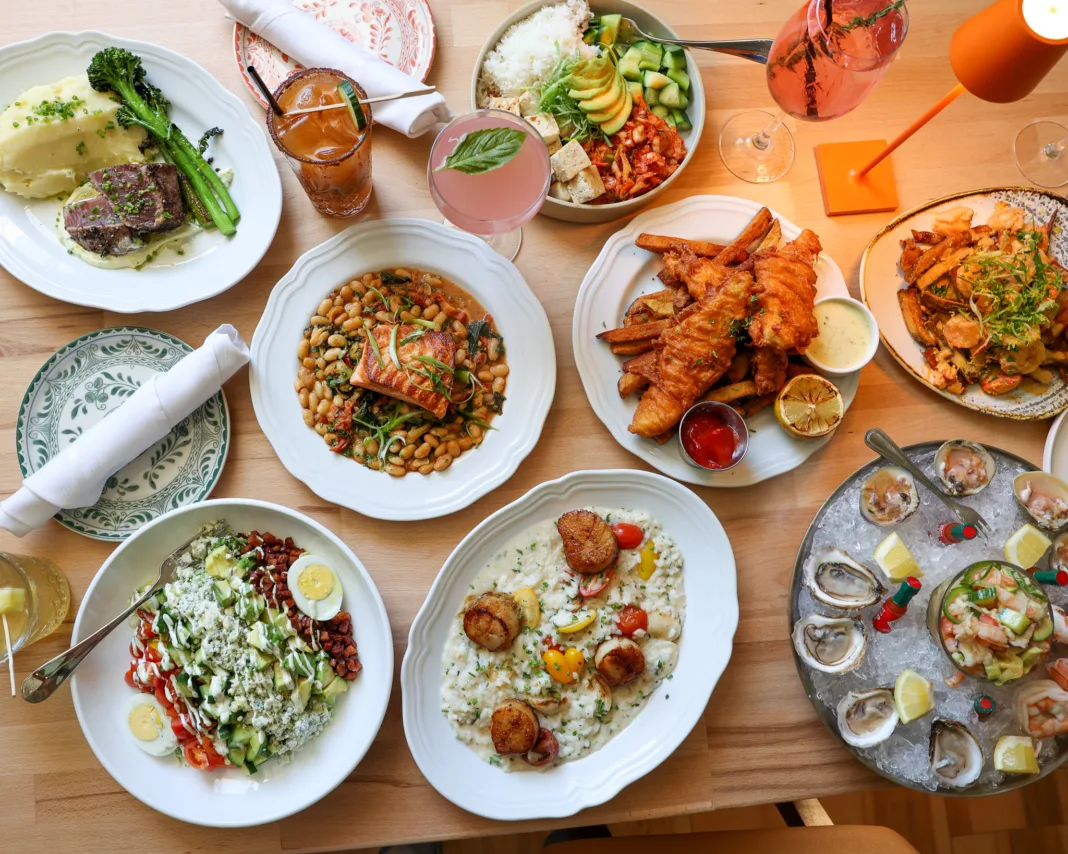 Overhead view of Coastal Canteen's seafood and American dining spread in Manasquan, New Jersey, featuring oysters, shrimp ceviche, scallop risotto, fish and chips, grilled salmon, steak, fresh salads, cocktails, and seasonal entrées