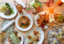 Overhead view of Coastal Canteen's seafood and American dining spread in Manasquan, New Jersey, featuring oysters, shrimp ceviche, scallop risotto, fish and chips, grilled salmon, steak, fresh salads, cocktails, and seasonal entrées