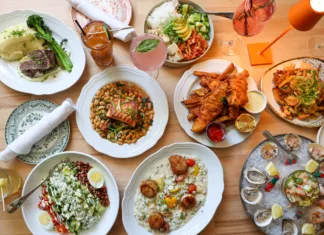 Overhead view of Coastal Canteen's seafood and American dining spread in Manasquan, New Jersey, featuring oysters, shrimp ceviche, scallop risotto, fish and chips, grilled salmon, steak, fresh salads, cocktails, and seasonal entrées