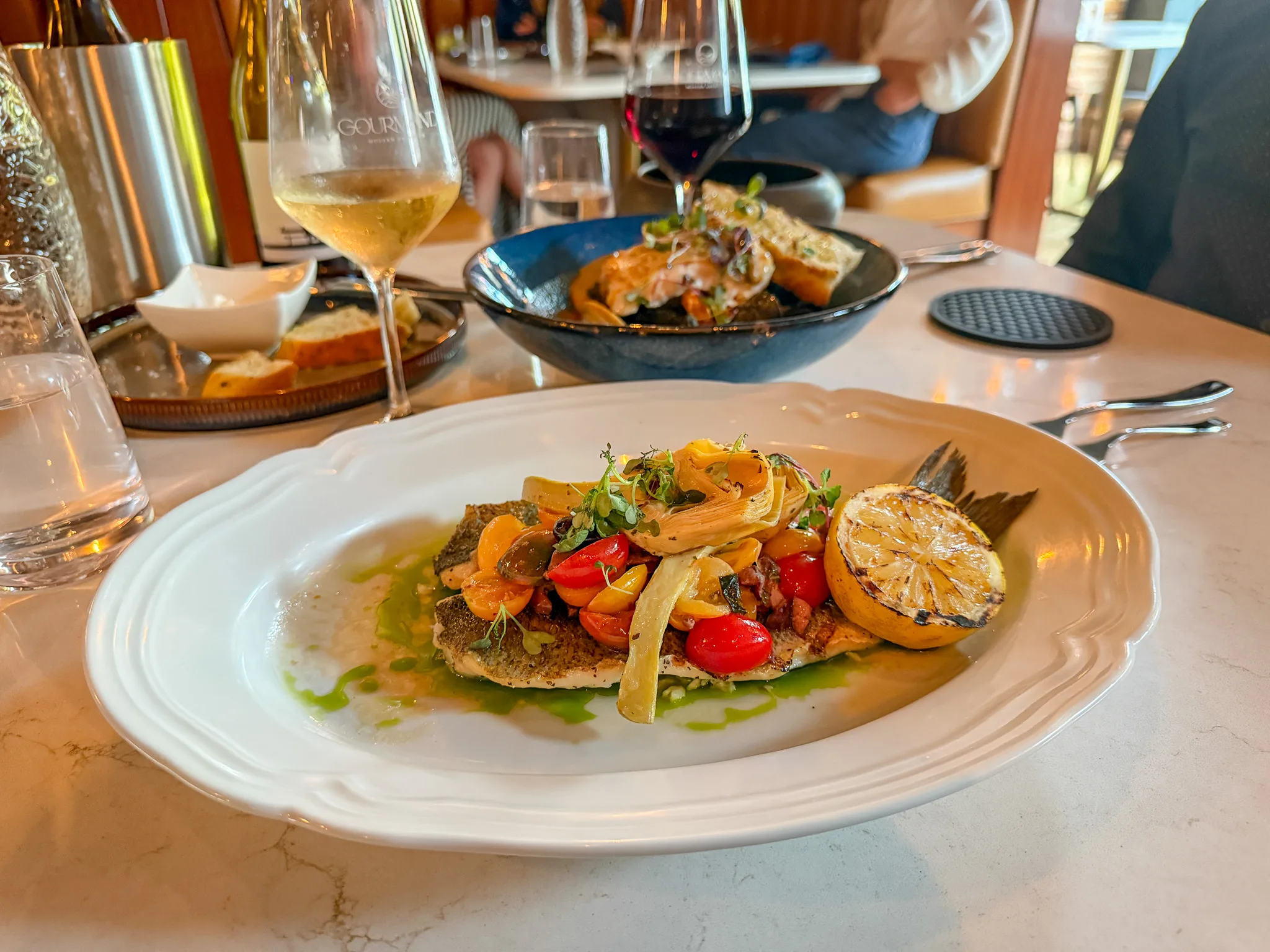 Pan-seared fish topped with roasted vegetables and grilled lemon, served at Gourmand restaurant in Point Pleasant Beach, New Jersey, with wine and upscale dining ambiance in the background