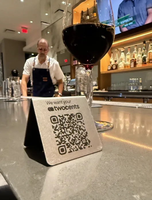 A twocents QR code tent card displayed on a restaurant bar invites guests to share private feedback with management.