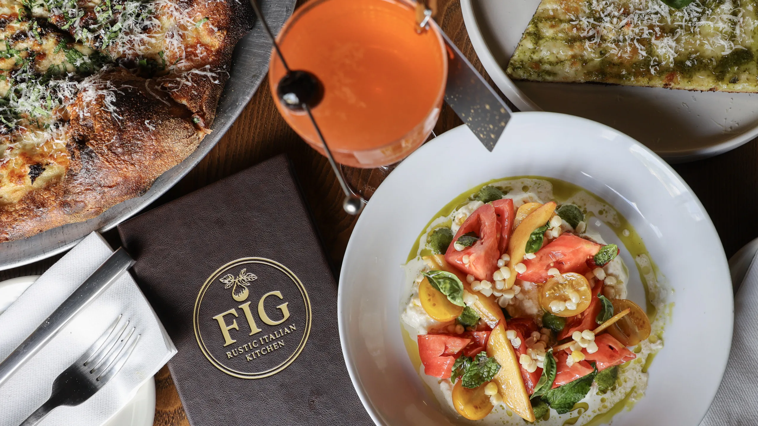 Table featuring Italian dishes, including a pizza, seasonal tomato and fruit salad, focaccia, and an orange cocktail, with a FIG Rustic Italian Kitchen menu.