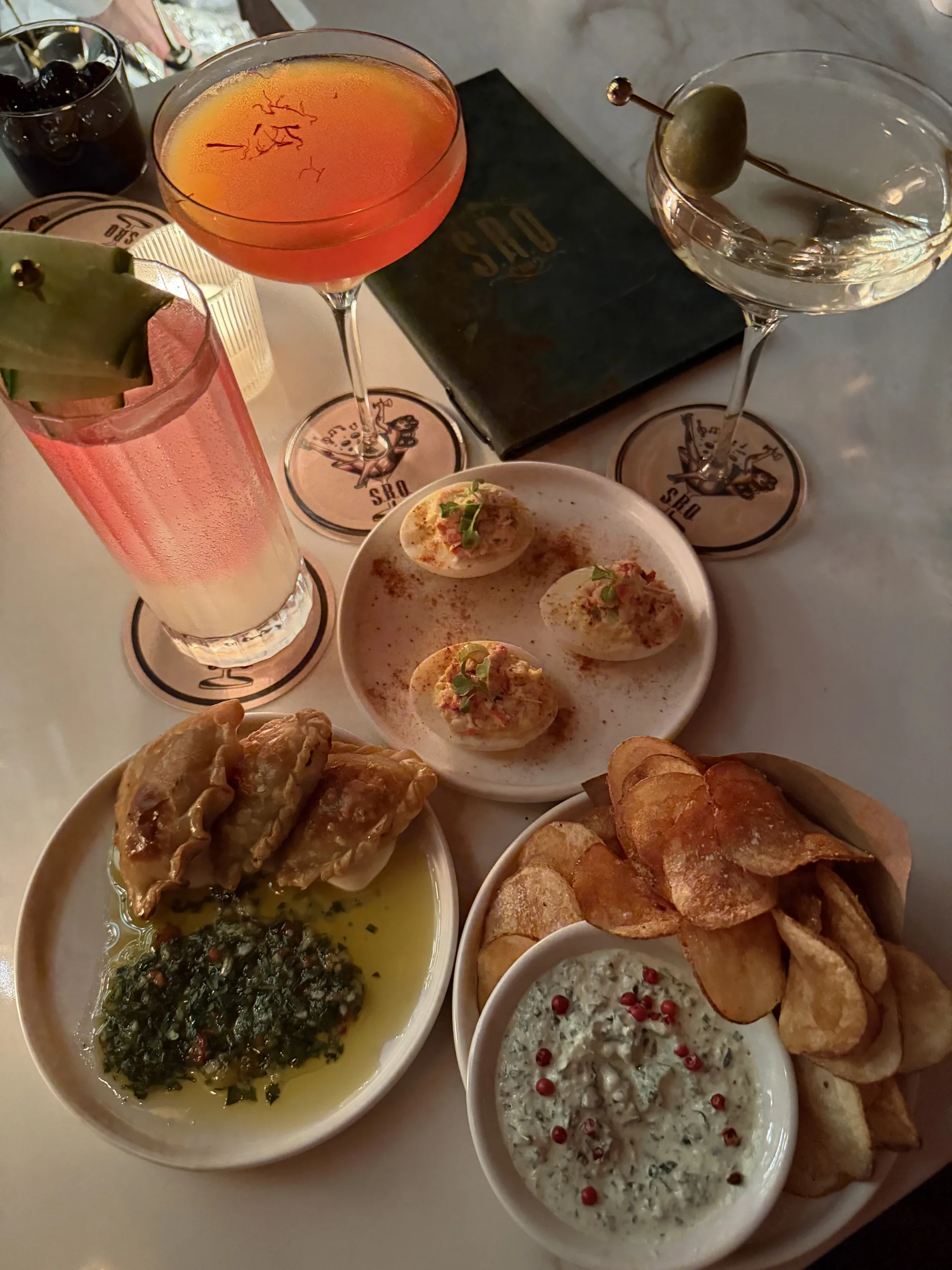 Assortment of cocktails and appetizers on a restaurant table, including deviled eggs, savory pastries with dipping sauce, potato chips, and a creamy dip.