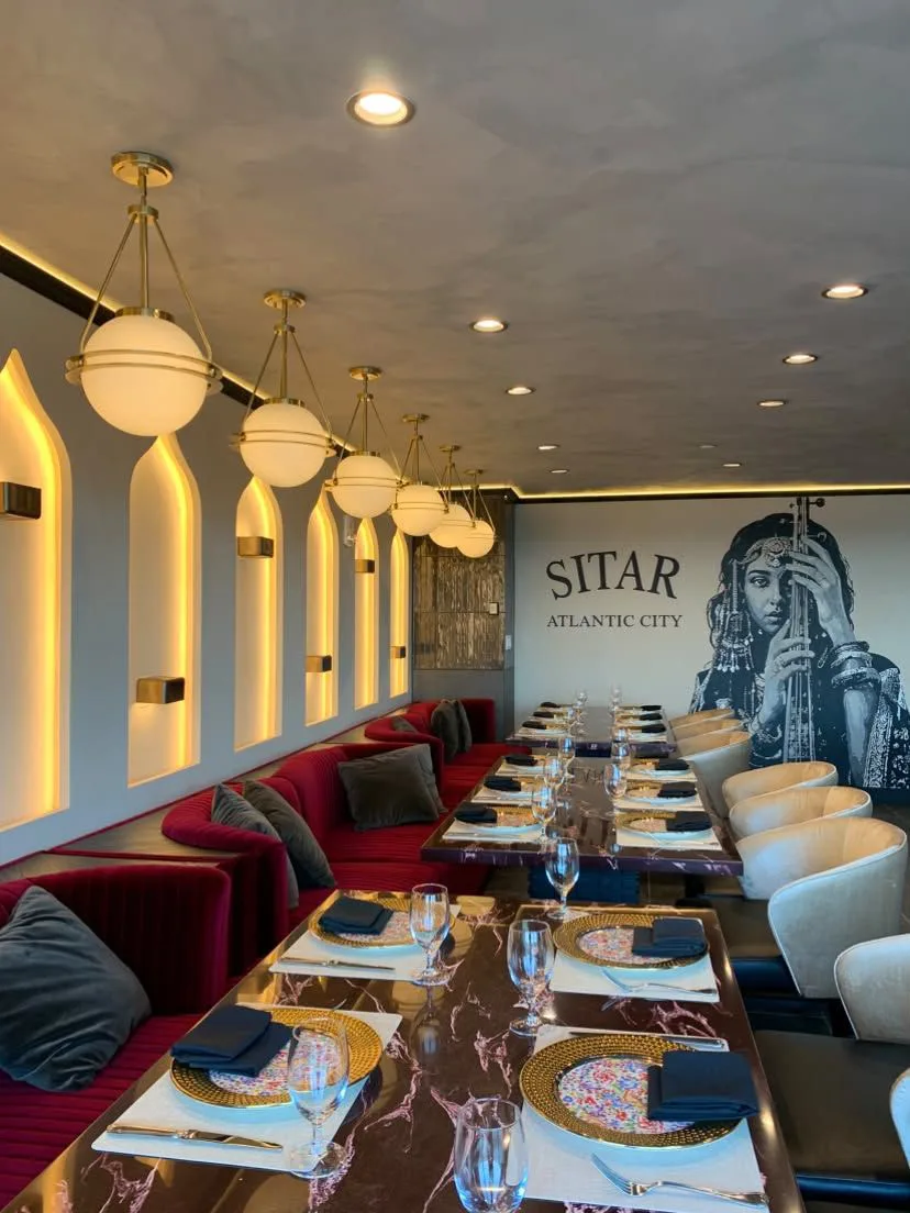 Elegant interior of Sitar Atlantic City featuring red velvet banquette seating, white chairs, marble tables set with colorful plates and glassware, gold globe pendant lights, and a large mural of a woman playing a stringed instrument.