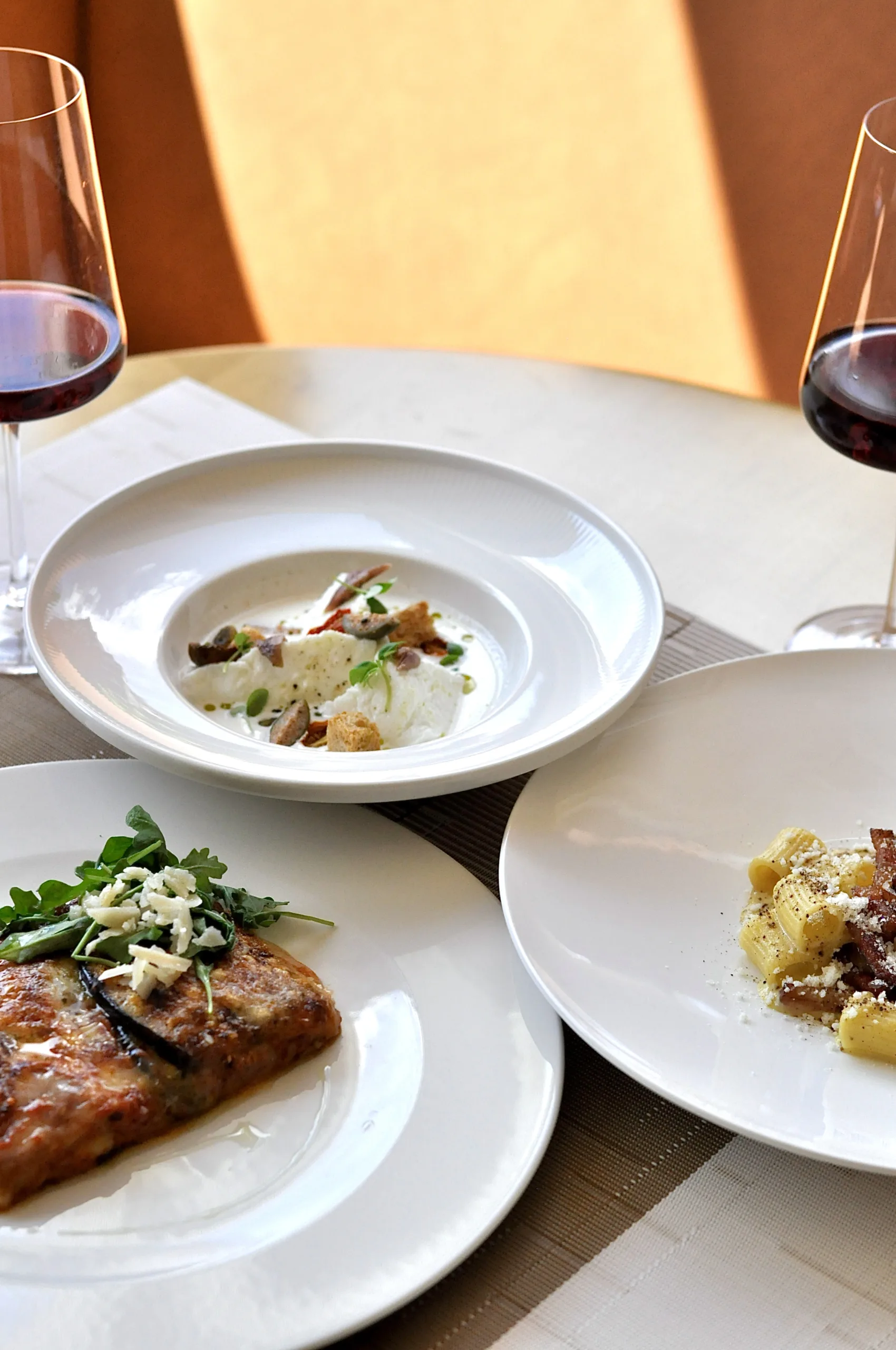 Three plates of Italian dishes at Clementina in Montclair, including a pasta dish, a plated entrée topped with greens and cheese, and a creamy appetizer, with two glasses of red wine on the table.