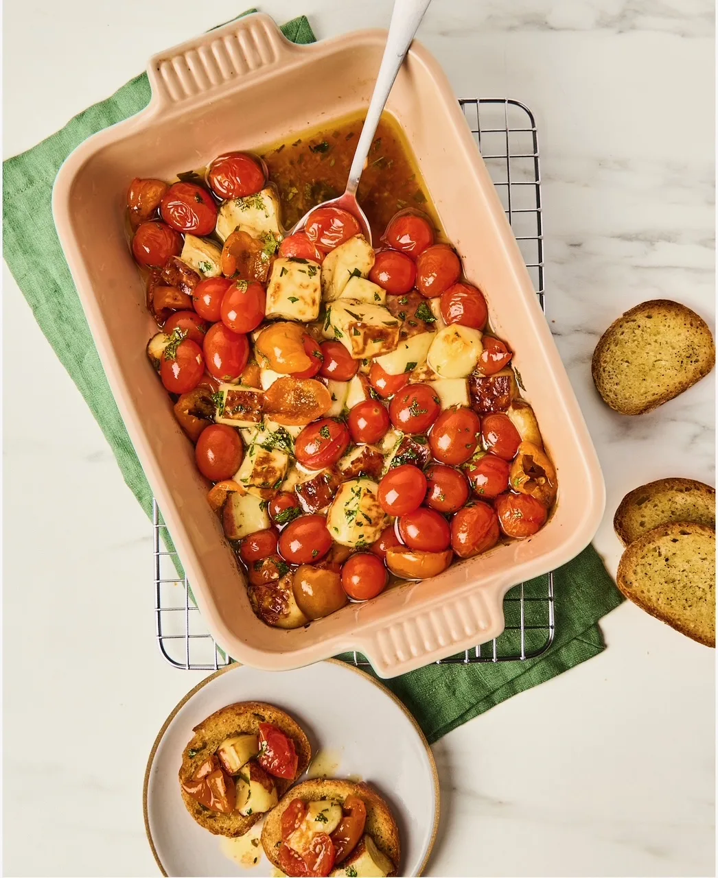 A baking dish filled with roasted cherry tomatoes, browned cheese, garlic, and herbs, served with toasted bread on the side.