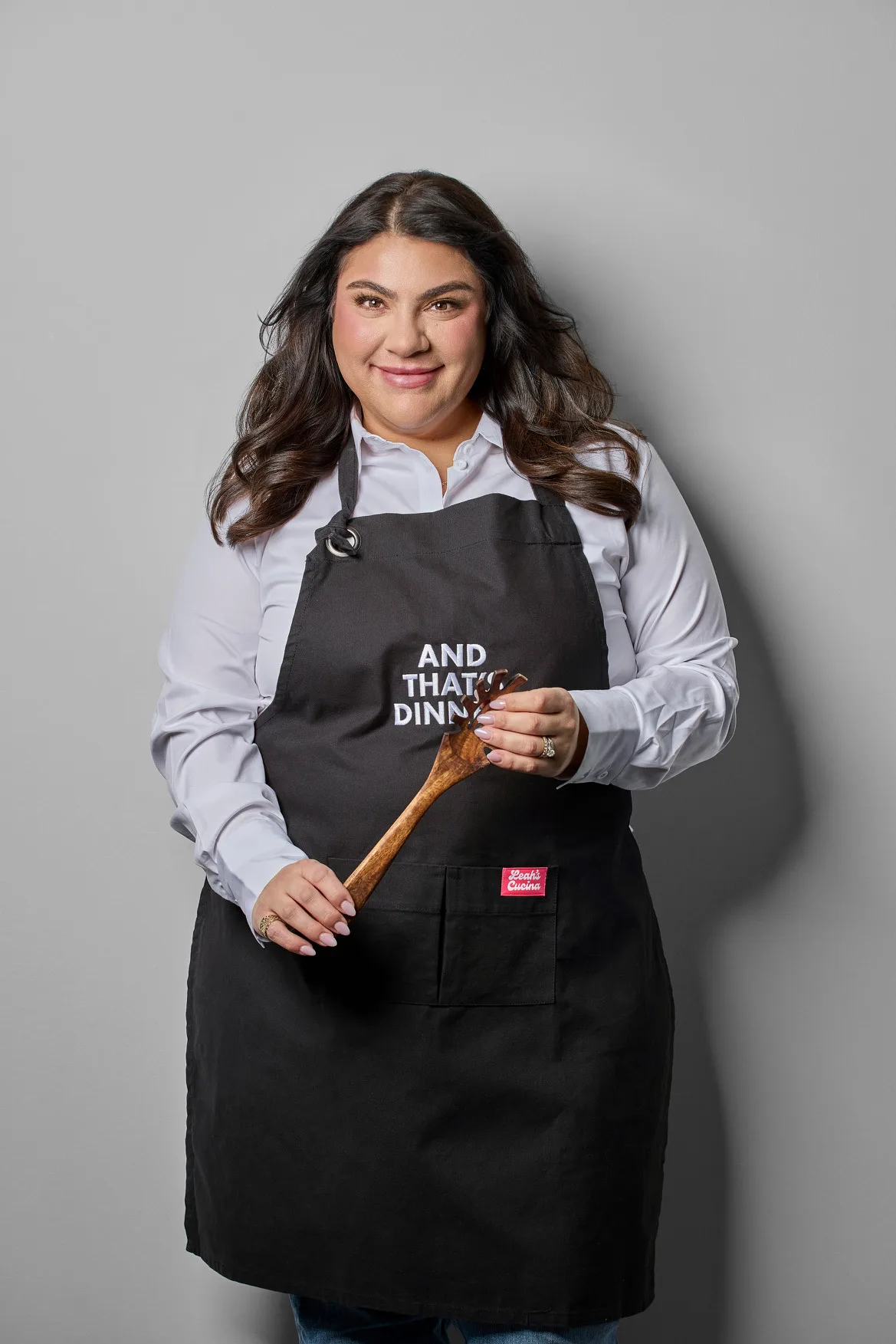 Portrait of cookbook author Leah Victoria wearing a black “And That’s Dinner!” apron over a white shirt and holding a wooden spoon against a gray background.