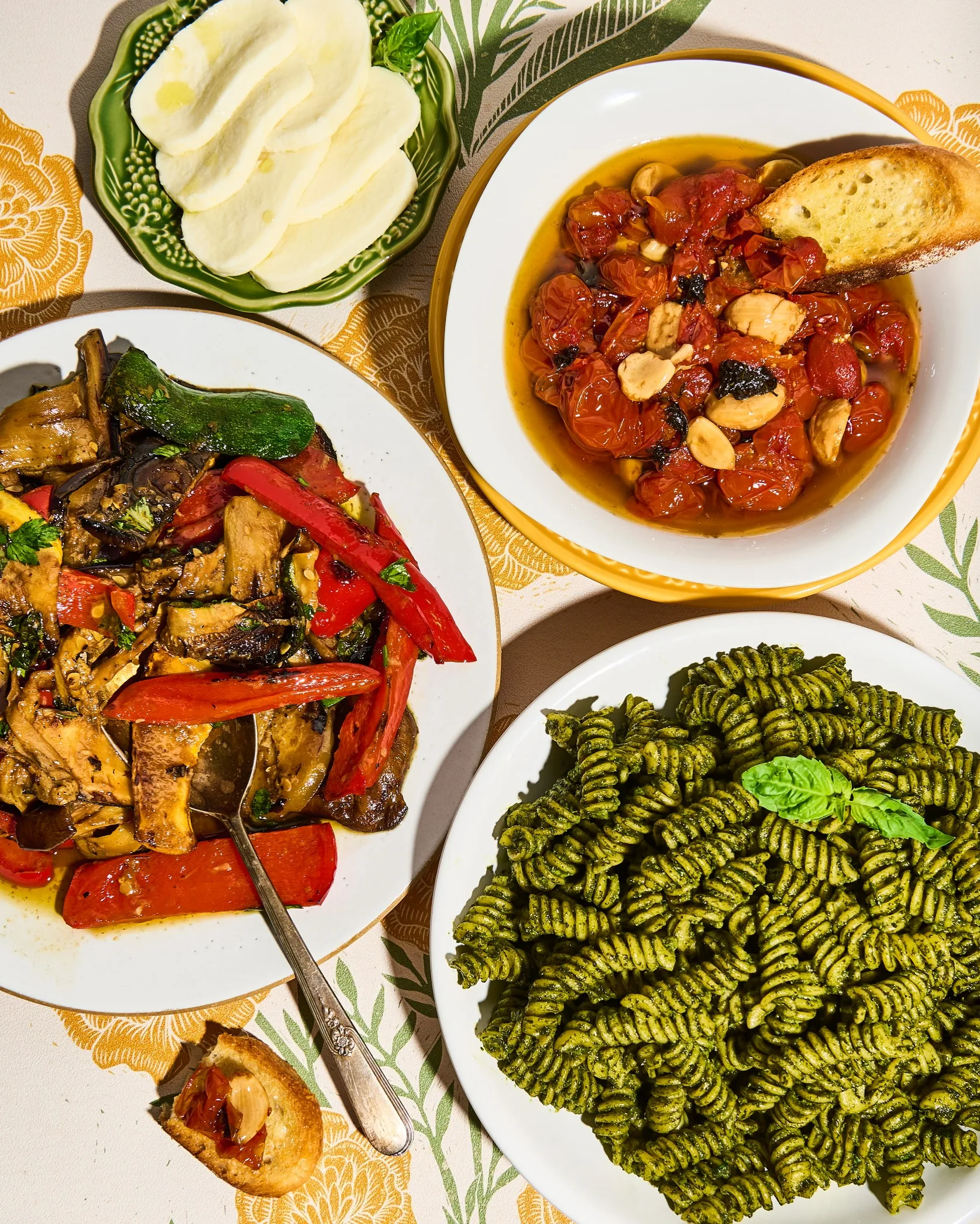 Three dishes from “And That’s Dinner!”: pesto rotini pasta, roasted mixed vegetables, and roasted tomatoes with garlic served with toasted bread.