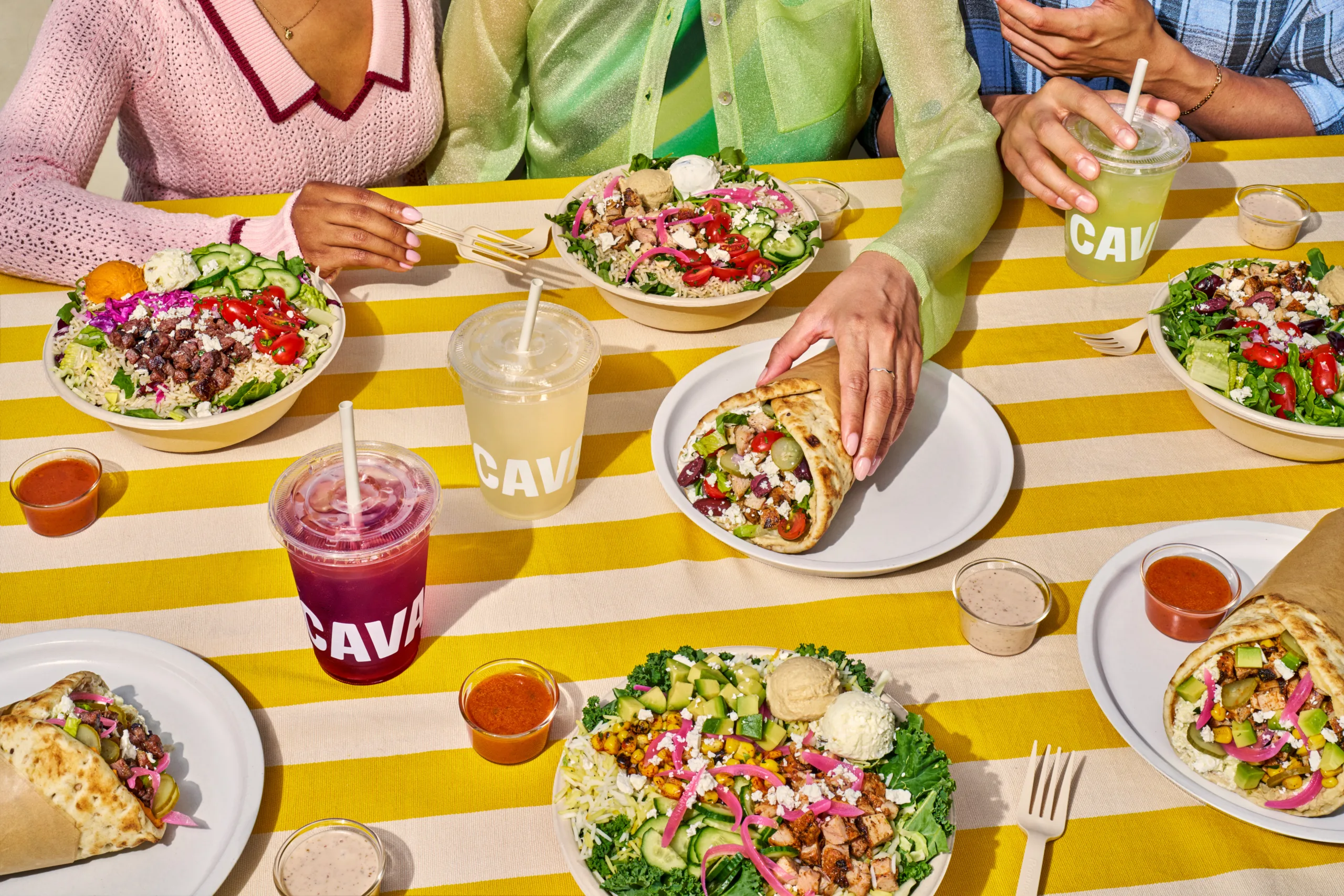 CAVA Mediterranean bowls, pita wraps, fresh salads, dips, and drinks spread across a yellow-striped table.