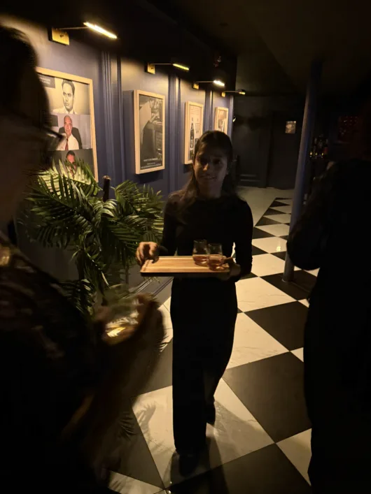 Server carrying whisky samples through the House of Amrut tasting room, featuring dark blue walls and black-and-white checkered flooring.
