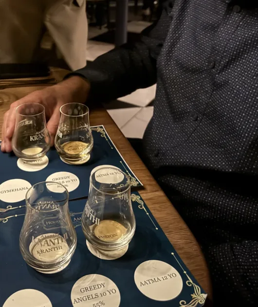 Amrut whisky tasting glasses arranged on a branded tasting mat with labels for Kranthi, Fusion, Greedy Angels, Gymkhana, and Aatma.