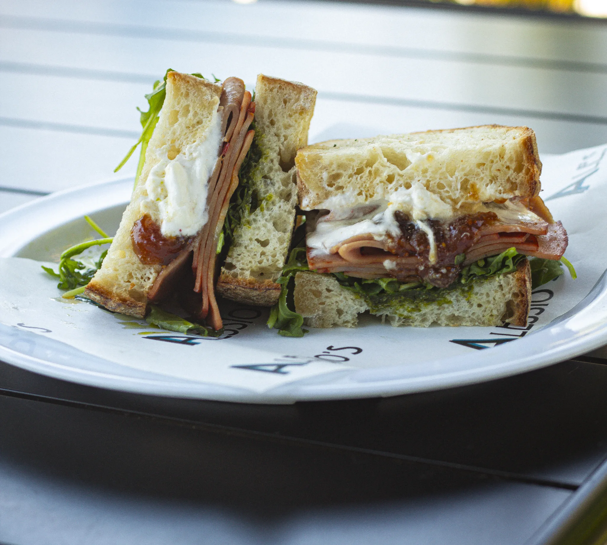 Sliced deli meat, fresh greens, and creamy white cheese layered between thick slices of toasted artisan bread on a branded plate.