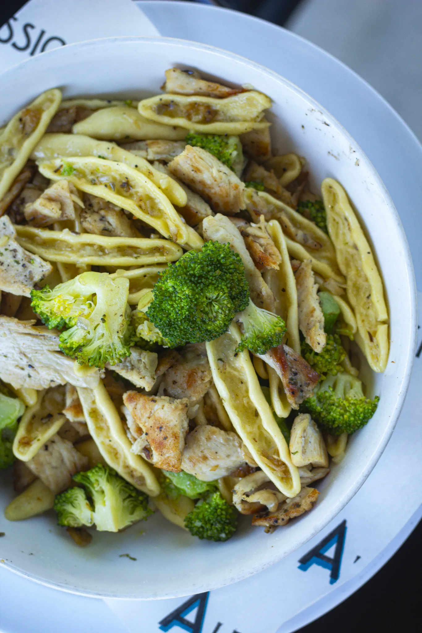 A bowl of pasta with grilled chicken, fresh broccoli, and a light sauce, served on a white restaurant plate.