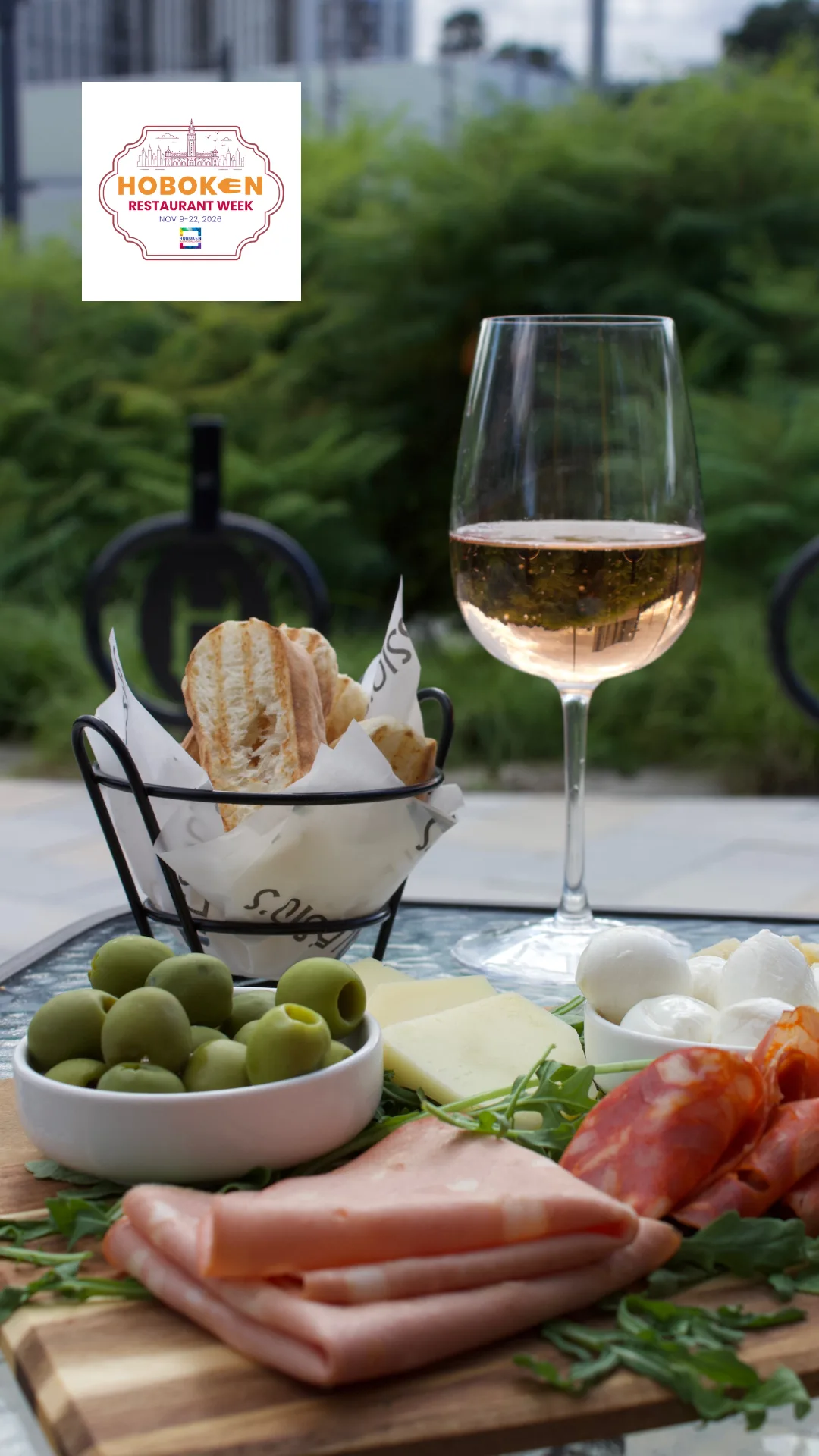 An Italian charcuterie spread featuring sliced deli meats, fresh mozzarella, cheese, green olives, arugula, and crusty bread, served with a glass of white wine. A Hoboken Restaurant Week graphic appears in the upper-left corner.